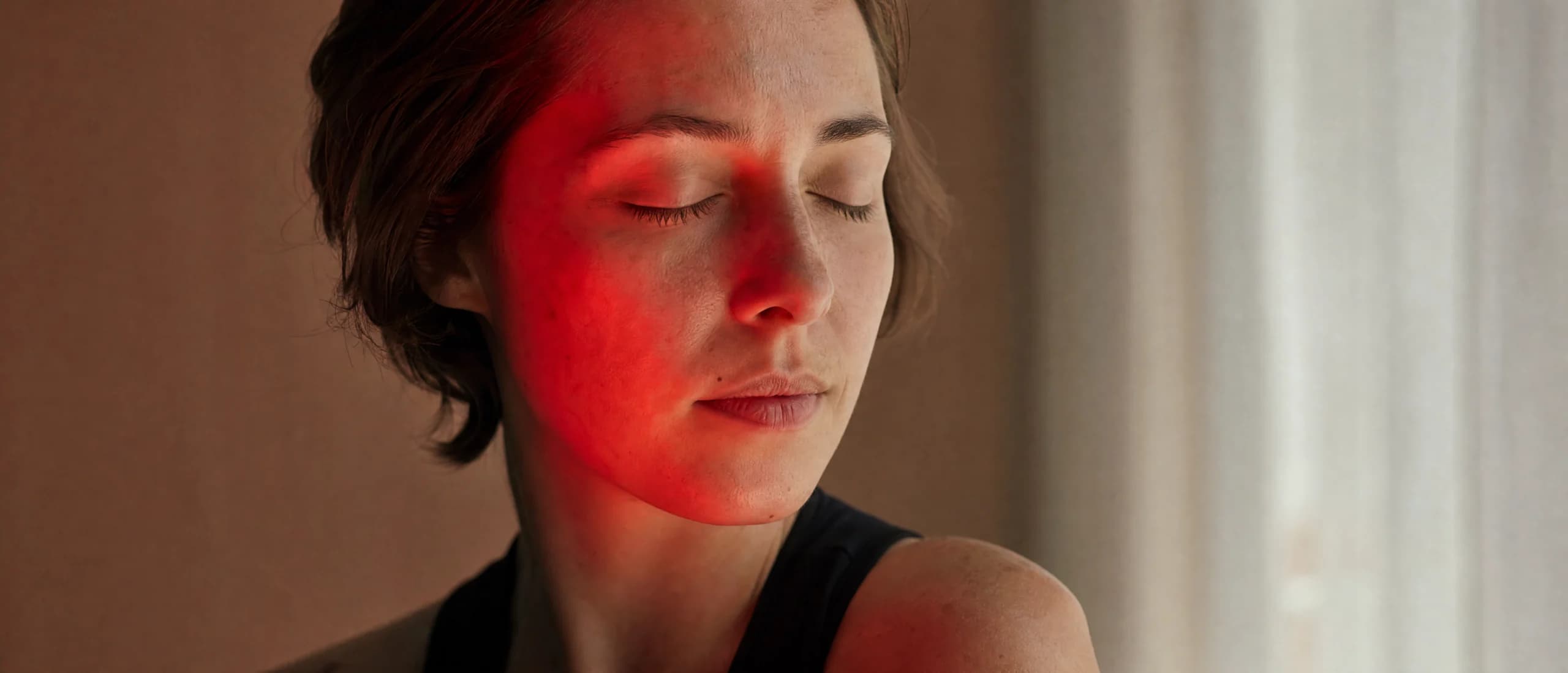 A woman with her eyes closed, one half of her face lit by a soft deep-red light, the other by daylight.