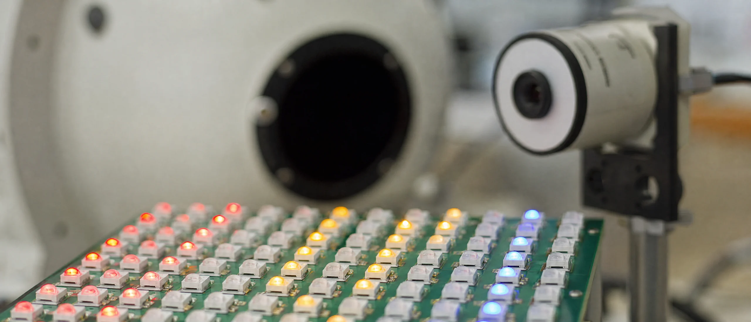 A dense array of small emitters on a circuit board, several lit in red, amber and blue, in front of an integrating sphere and a detector head.